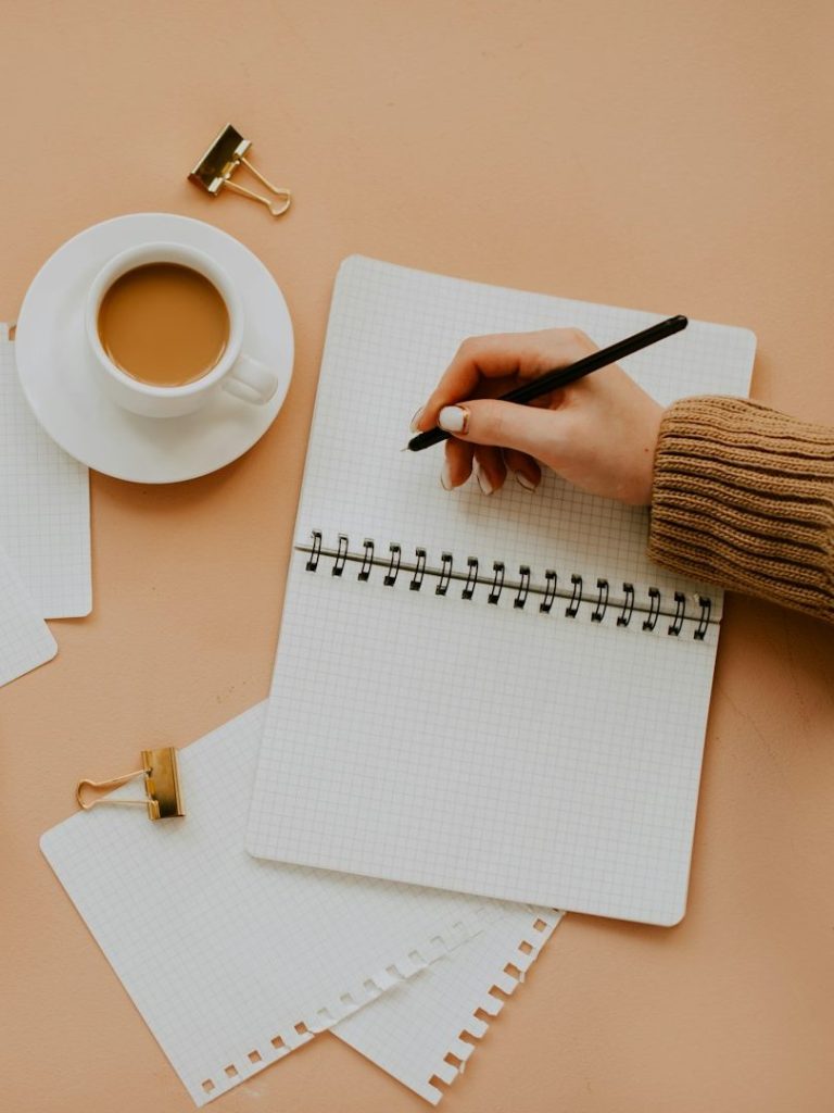 Main en train d'écrire dans un carnet avec une tasse de café et des feuilles blanches.